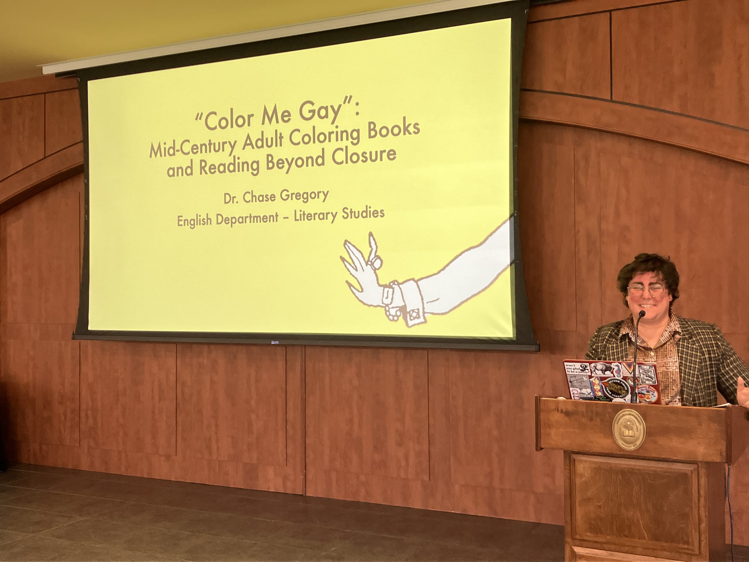 Chase Gregory at a lectern with PowerPoint title page behind them. The title of the presentation is "Color Me Gay: Mid-Century Adult Coloring Books and Reading Beyond Closure."